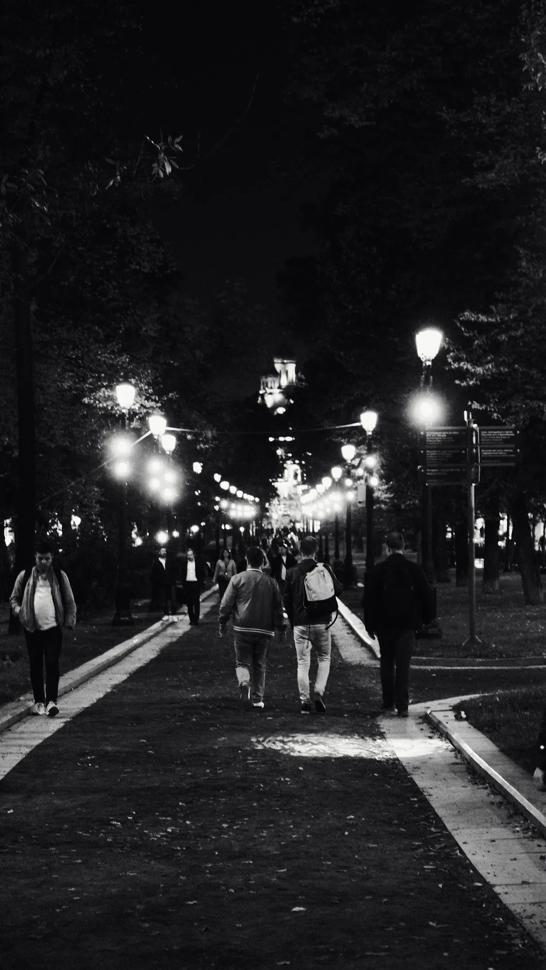 Paseo peatonal iluminado por farolas, en blanco y negro, con personas caminando.
